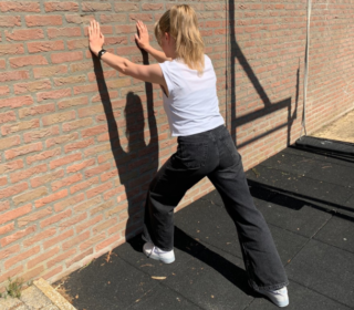 Een vrouw met een wit shirt en zwarte spijkerbroek is een oefening tegen de muur aan het doen